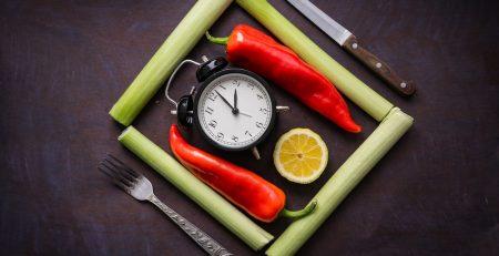 Place setting with a old style ringing alarm clock in the middle and a slice of orange. Flanked by 2 red chili peppers, surrounded in a square by 4 celery stalks. A fork and knife on the outside.