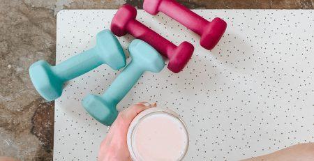 Overhead view of a person sitting cross legged holding a drink of a creamy pink liquid. some light foam wrapped dumbbells' in teal and magenta are on the floor.