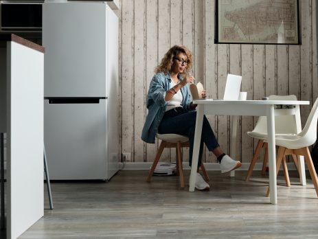 Lady sitting at a white table in front of a white laptop eating with chopsticks out of a take out container