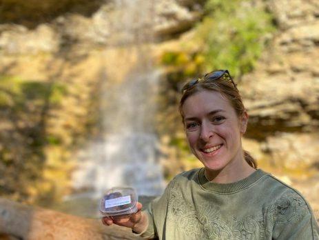 Hiker in front of a blurry waterfall. Holding a small container of food with a label on the top