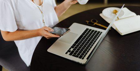 Lady holding a phone in one hand, tea mug in the other seated in front of a laptop