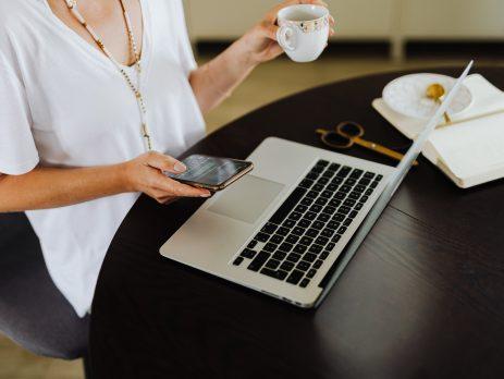 Lady holding a phone in one hand, tea mug in the other seated in front of a laptop