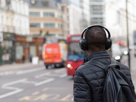 A person wearing headphones and a black jacket with a backpack standing on the sidewalk of a city